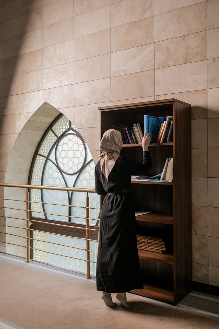 A woman in hijab chooses a book from a wooden shelf in a stylish indoor setting.