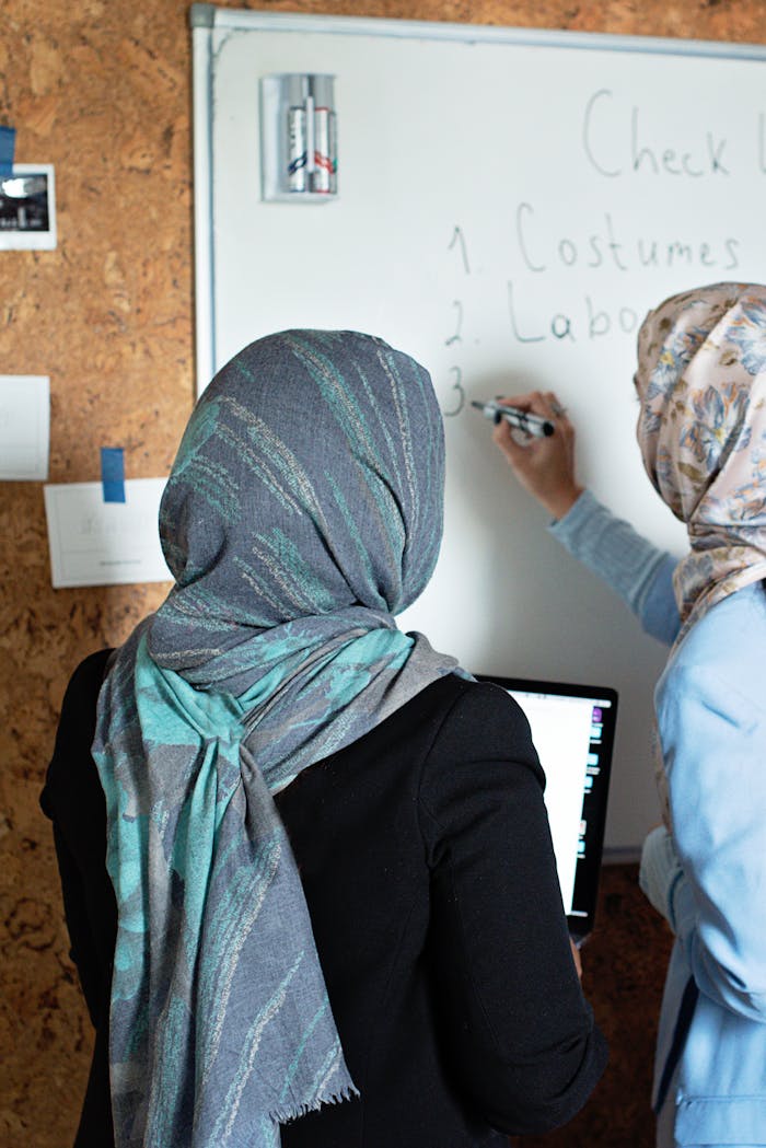 Two Muslim women wearing hijabs collaborate at a whiteboard in an office setting.